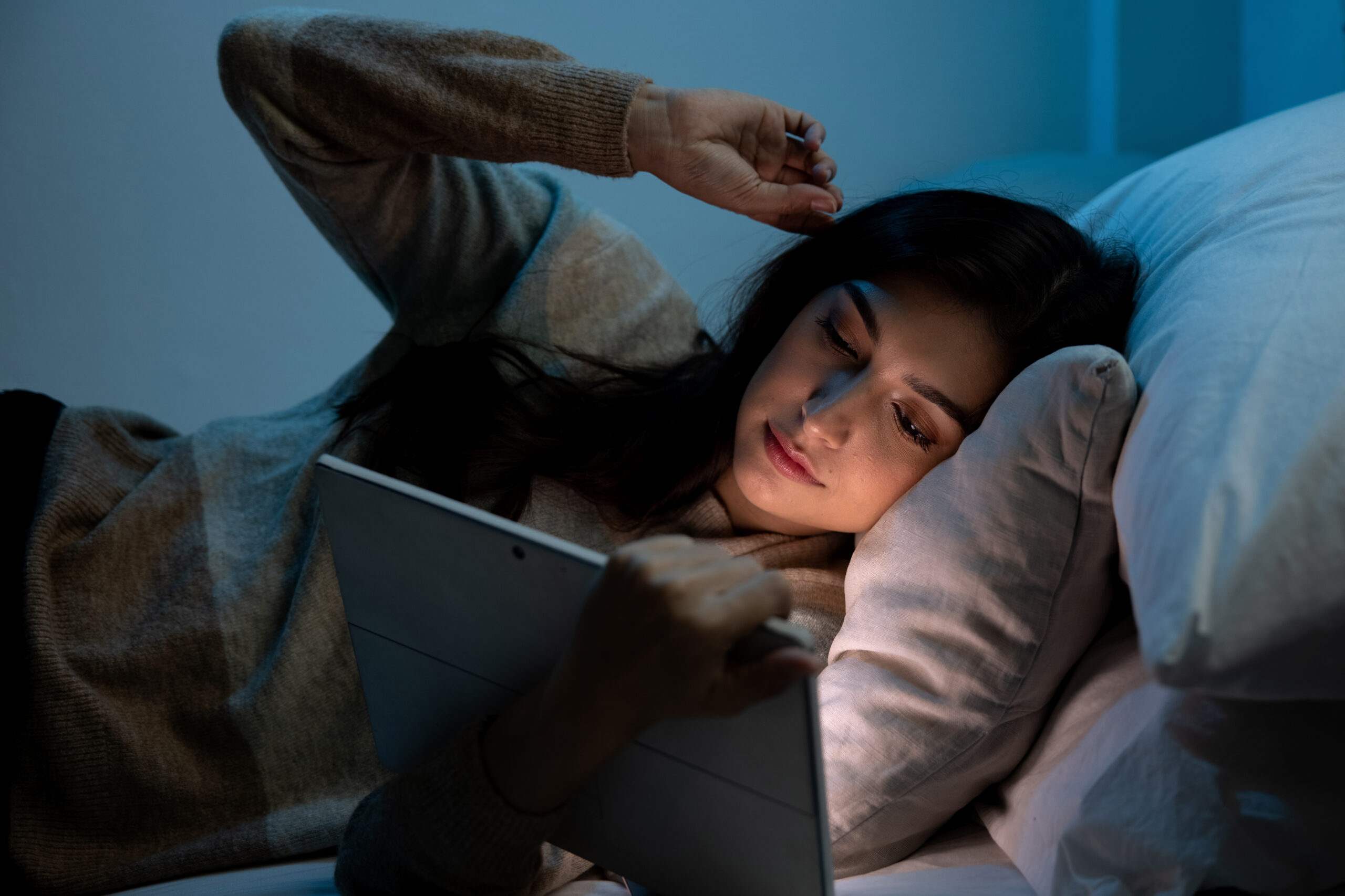 A woman lies in bed at night, using a tablet, highlighting the effects of blue light on sleep and ways to avoid it.