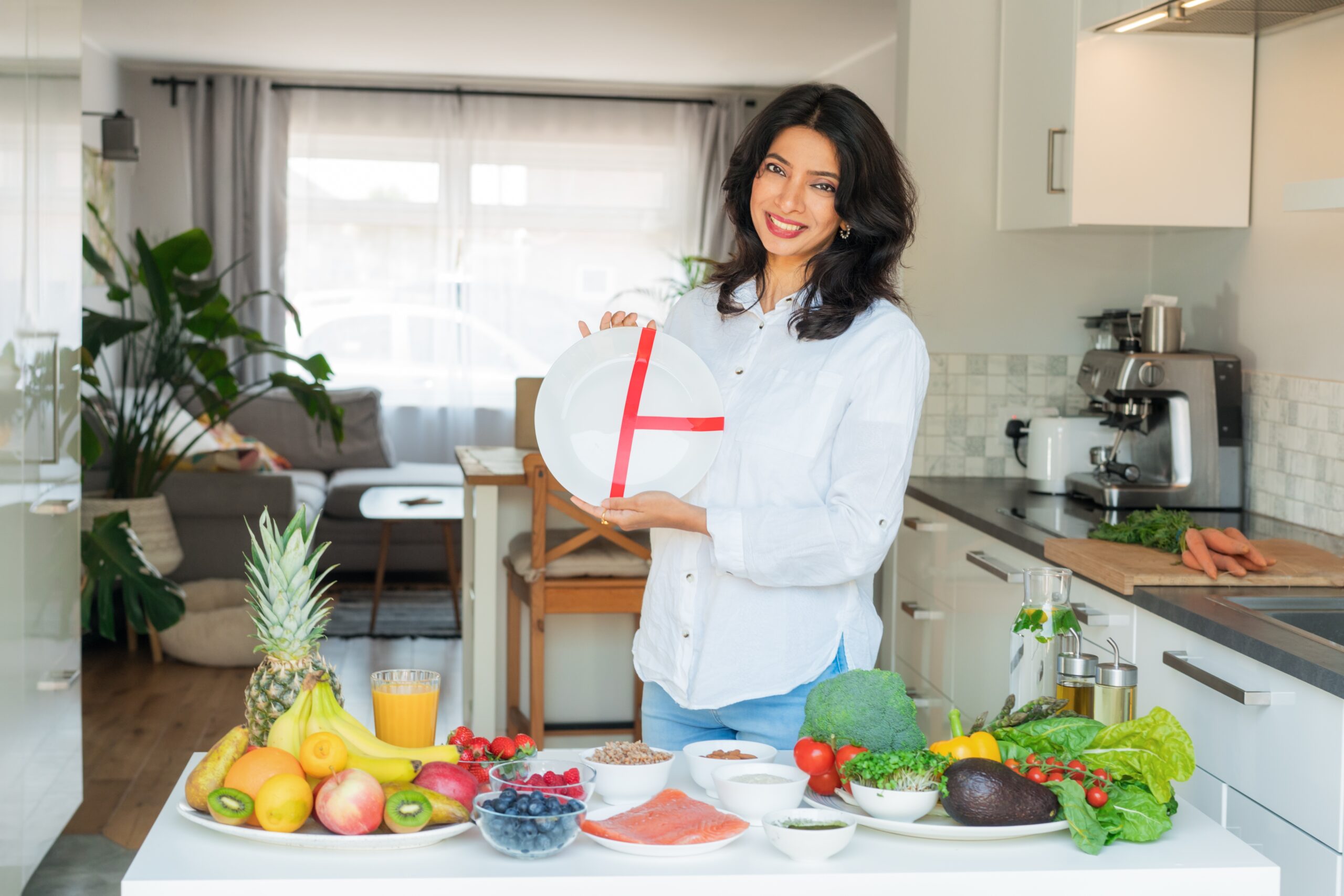 A woman smiles and points to a plate with markings, surrounded by fresh fruits and vegetables, illustrating the art of a balanced plate without dieting.