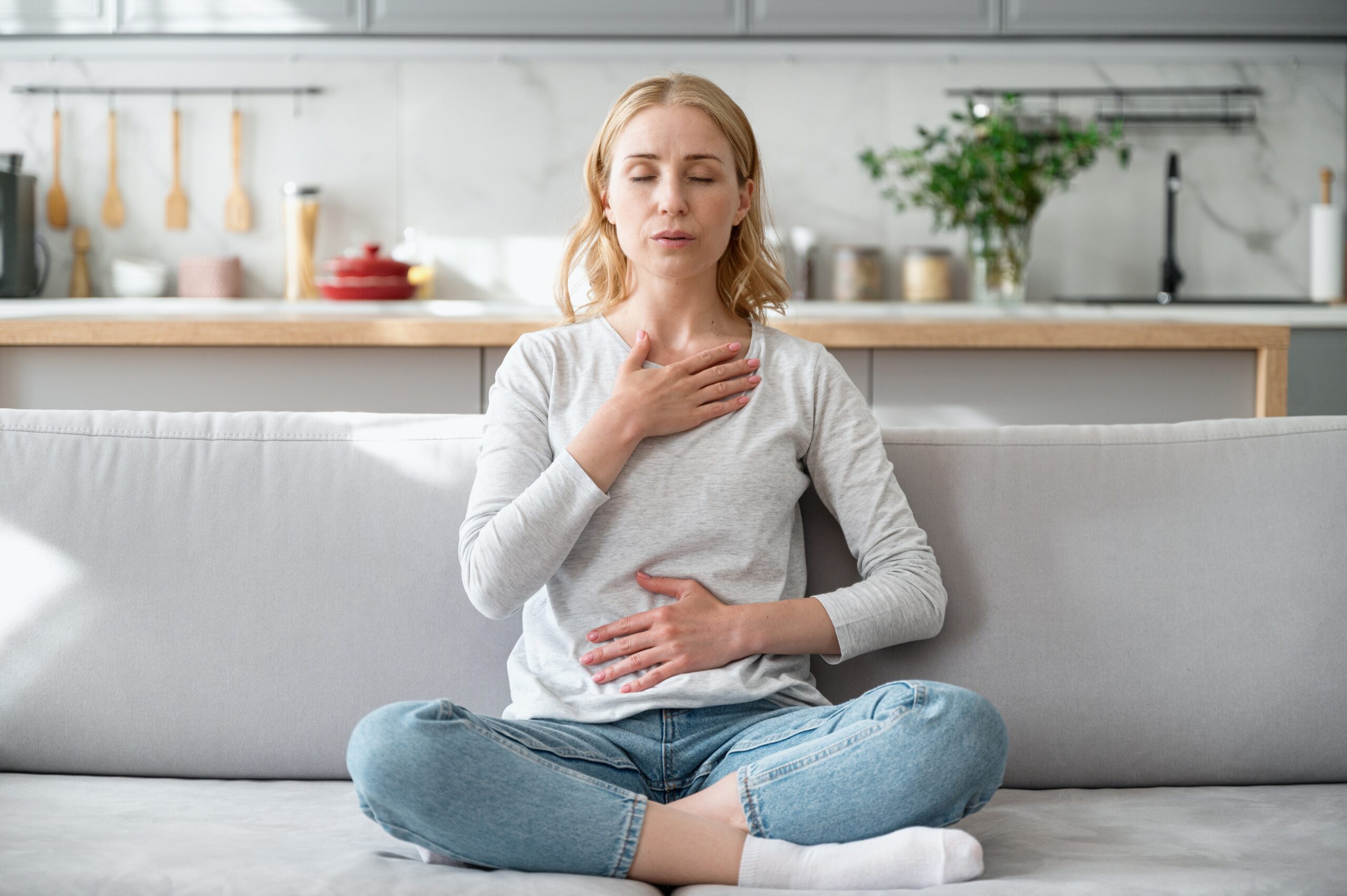 A woman sits comfortably on the sofa, eyes closed, one hand on her chest and the other on her belly, practicing breathing exercises to boost energy.