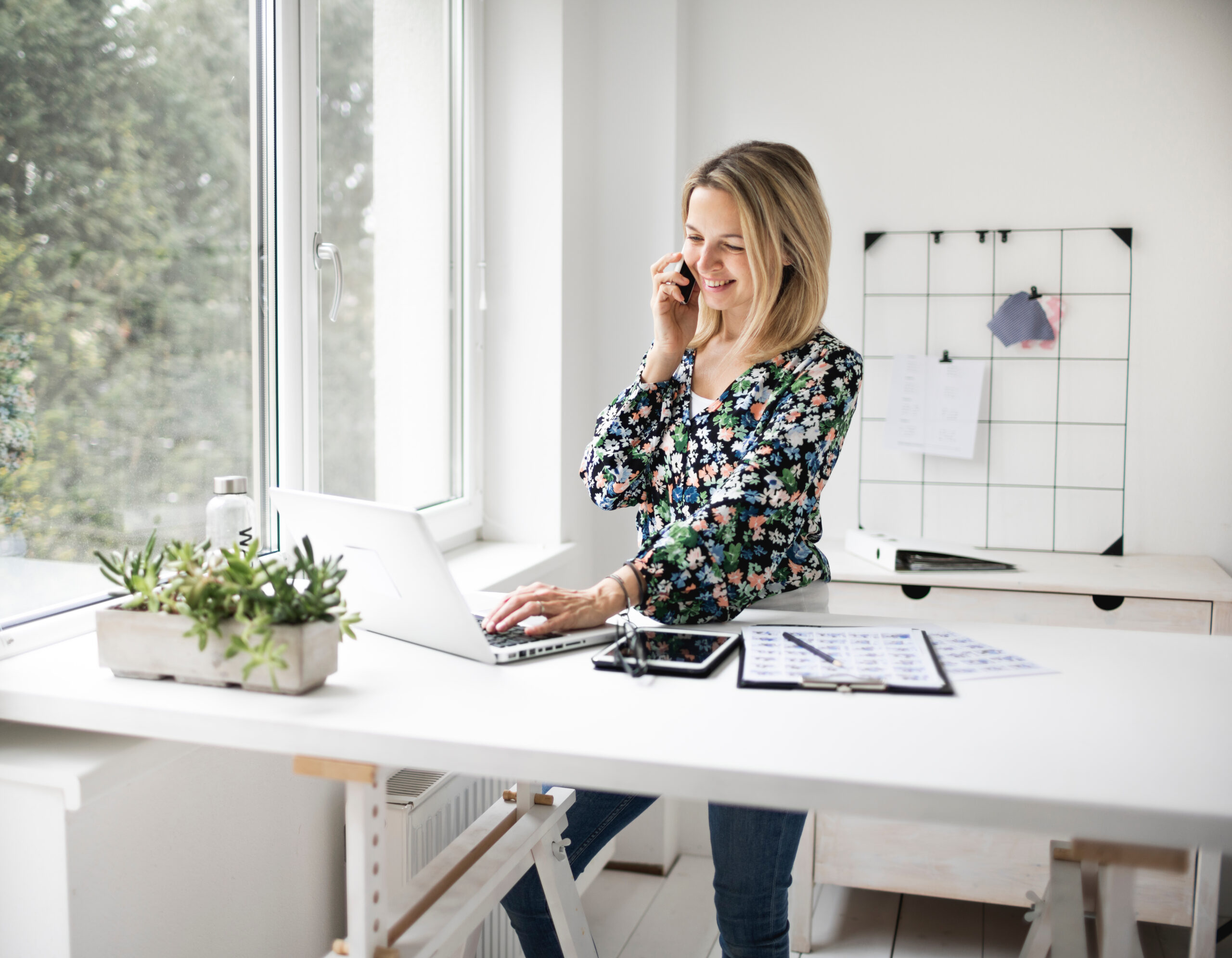 A woman stands smiling at her desk, talking on the phone and working on her laptop, highlighting the benefits of standing more and sitting less.