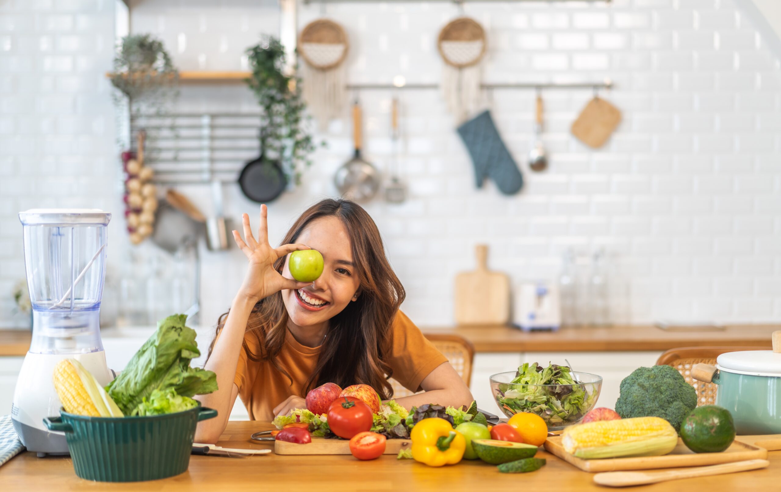 A woman playfully holds a green apple in front of her eye, surrounded by fresh fruits and vegetables, demonstrating how to simplify nutrition using the 80/20 principle.