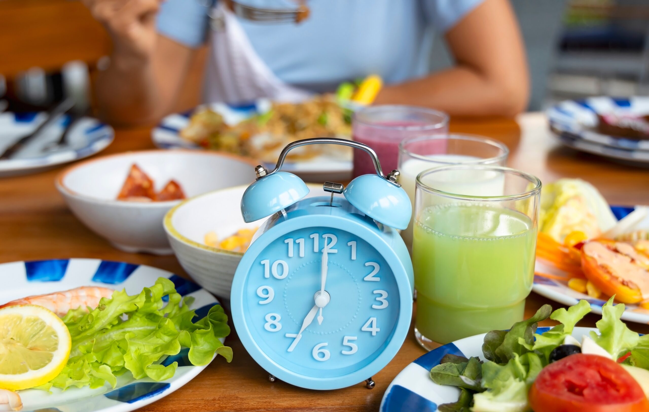 A blue alarm clock sits among plates of food, symbolizing the timing of meals in intermittent fasting.